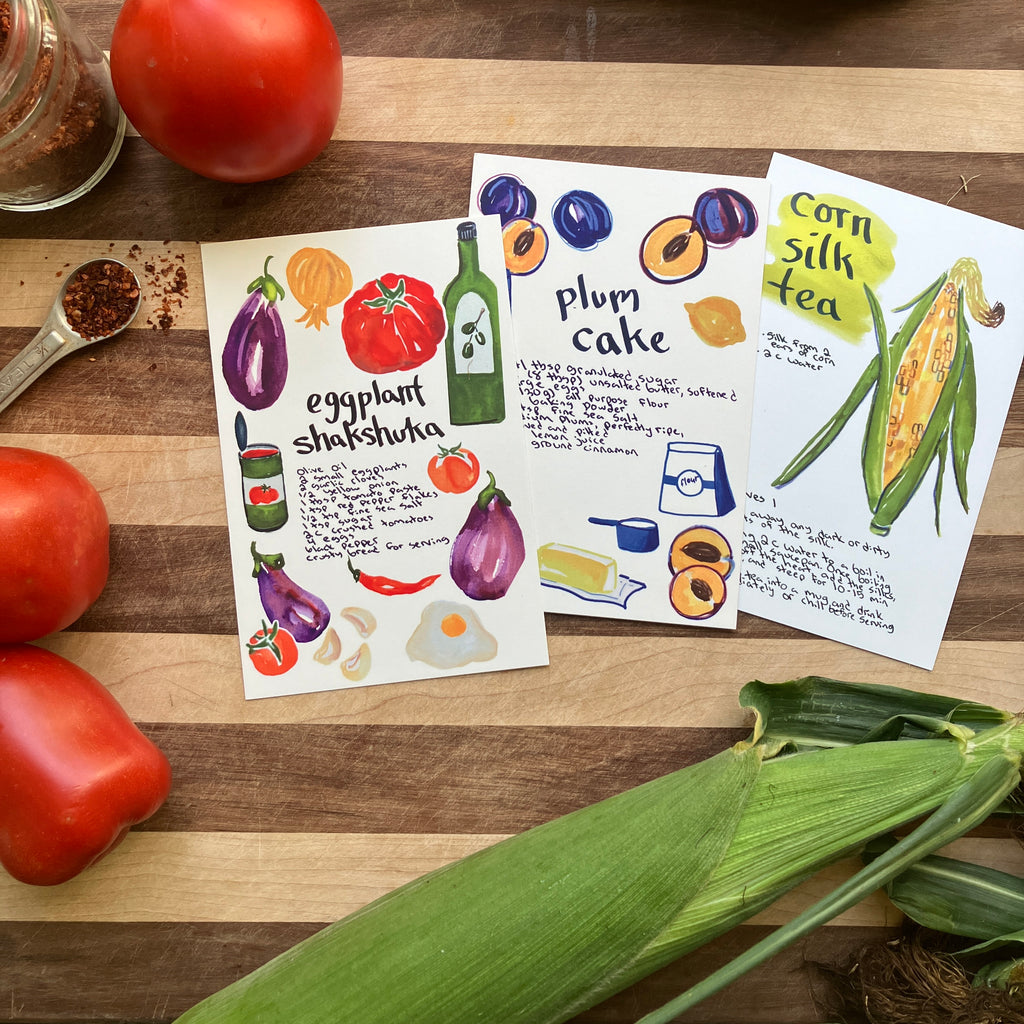 Three illustrated recipe cards laid on a wooden surface with tomatoes, a spoon of spices, and corn in the scene. The cards show hand-drawn recipes including eggplant shakshuka, plum cake, and corn silk tea.